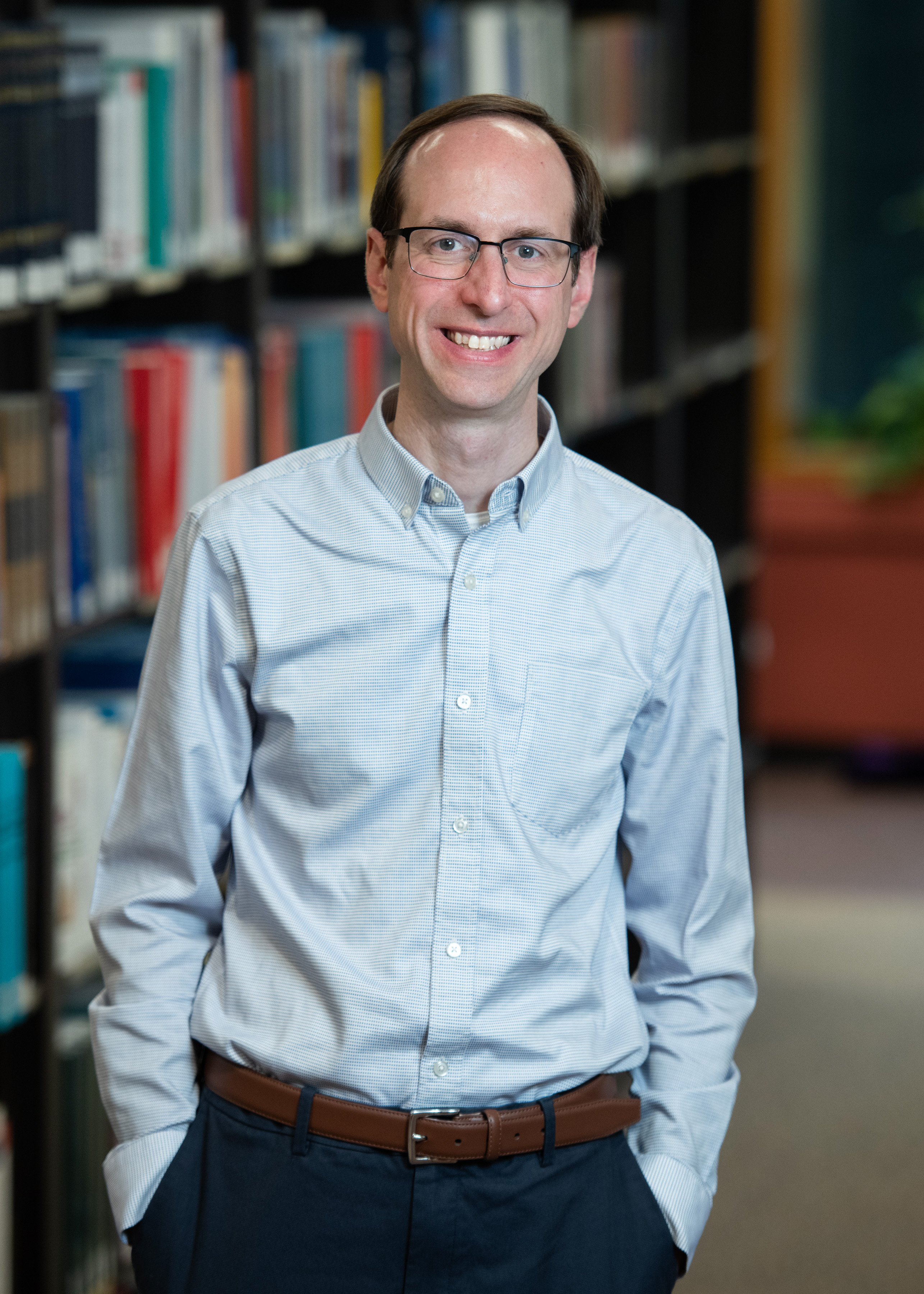 Matthew Richardson, a white man in a pale blue collared shirt, poses and smiles in front of shelved books.