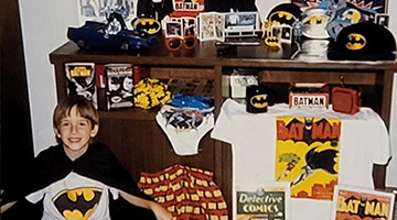 A smiling child in a Batman cape surrounded by other Batman merchandise. The photo is clearly aged.
