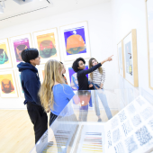 A group standing in front of mounted paintings. One of the group is gesturing at the painting and speaking. 