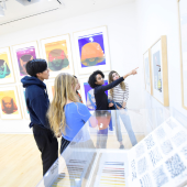 A group standing in front of mounted paintings. One of the group is gesturing at the painting and speaking