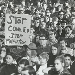 A black and white photograph of a large group of people, packed together. One holds up a sign, reading STOP COMM ED STOP POLLUTION