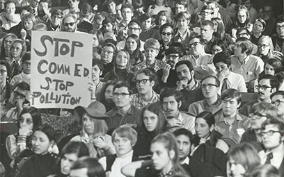 A black and white photograph of a large group of people, packed together. One holds up a sign, reading STOP COMM ED STOP POLLUTION